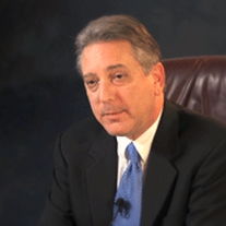 Portrait of a suited man seated in a brown leather chair on a gray background